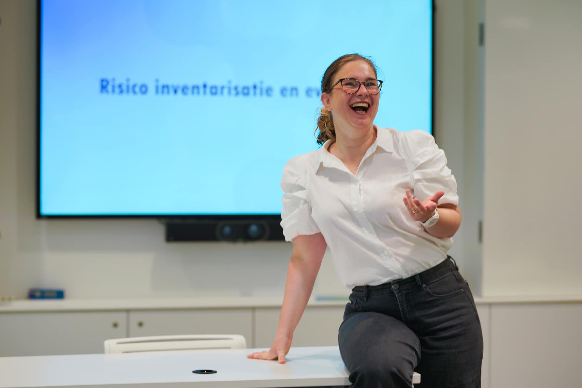 De vrouw op de foto zit met haar elleboog op een tafel en lacht. Ze draagt een wit overhemd met pofmouwen en een donkere broek. Haar bril heeft een opvallend montuur. Op de achtergrond is een blauw scherm te zien met de tekst