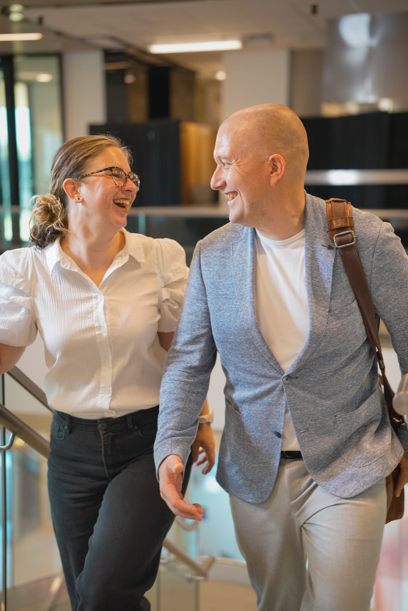 Een kale man met een blauw jasje en een vrouw met een bril en een wit overhemd lopen lachend samen een trap op. De man draagt een bruine schoudertas.