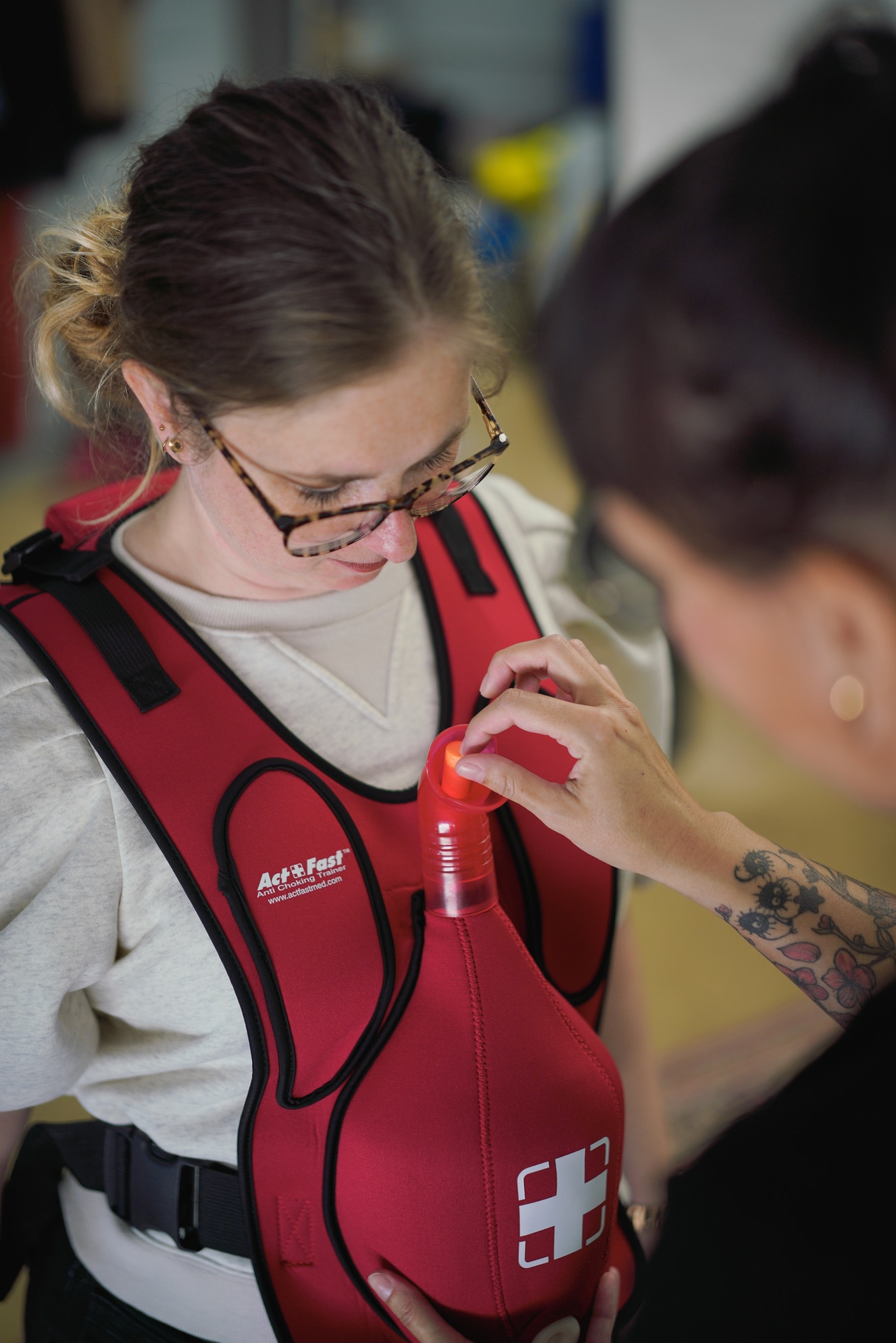 Een persoon draagt een rood vest met een wit medisch kruis erop, en kijkt naar beneden. Een ander persoon met tatoeages op de armen is bezig met het verstellen van het vest bij de borst.