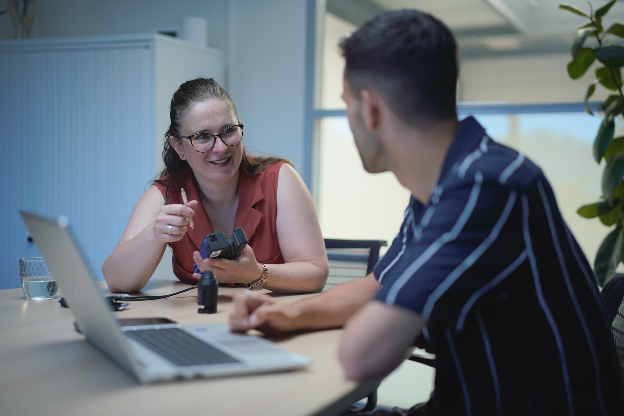 Een jonge vrouw zit aan een tafel met een man tegenover haar. Ze draagt een roodbruine top en een bril. De vrouw lacht naar de man terwijl ze een klein, elektronisch apparaat in haar handen heeft. Er staat een laptop en een glas water op de tafel. De scène vindt plaats in een kantooromgeving.