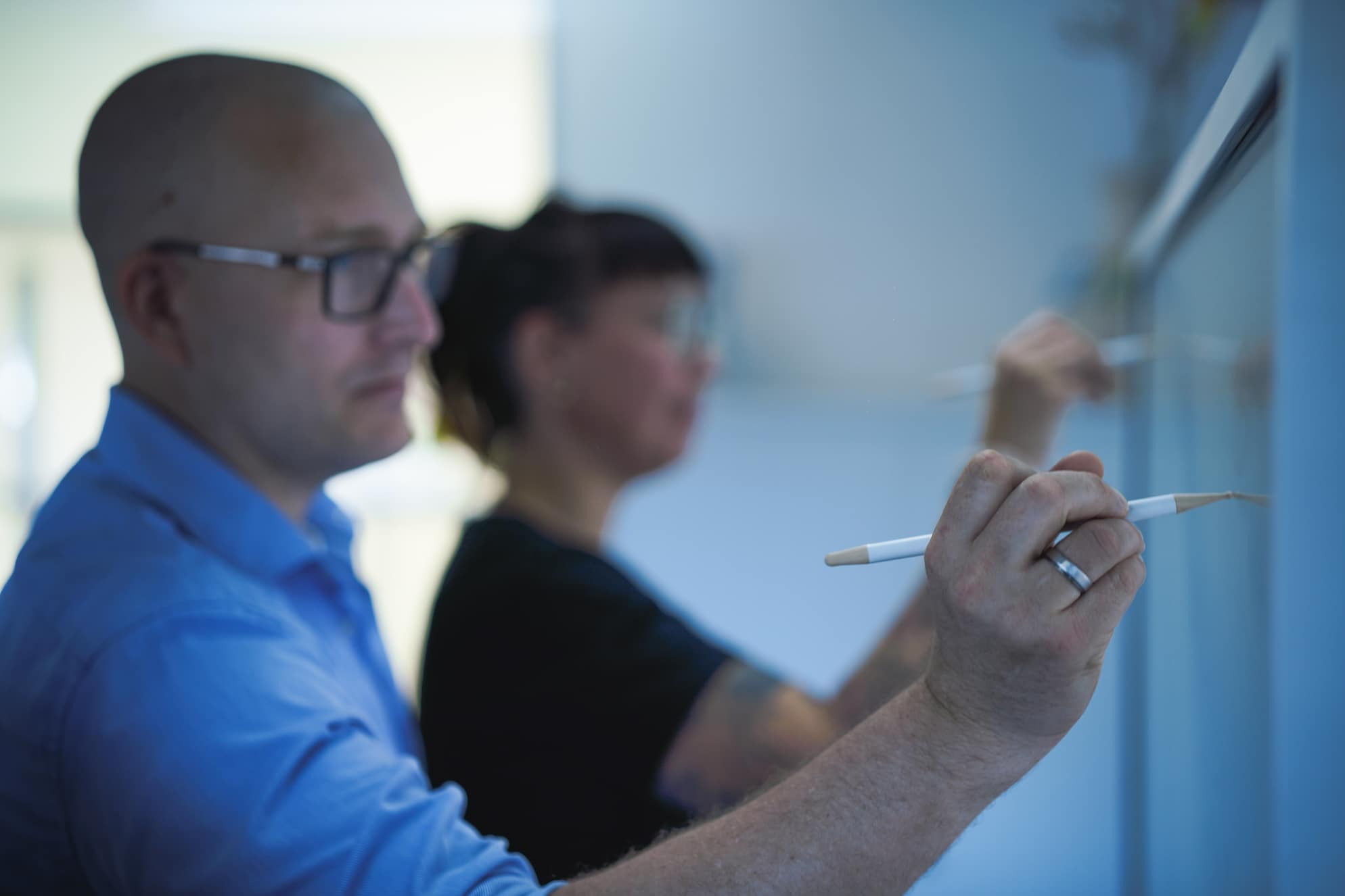 Twee personen staan naast elkaar en schrijven op een whiteboard. De focus ligt op de kale man op de voorgrond die een blauw overhemd en een bril draagt. De man houdt een stift vast. Op de achtergrond is een tweede persoon met donker haar en een bril vaag te zien.