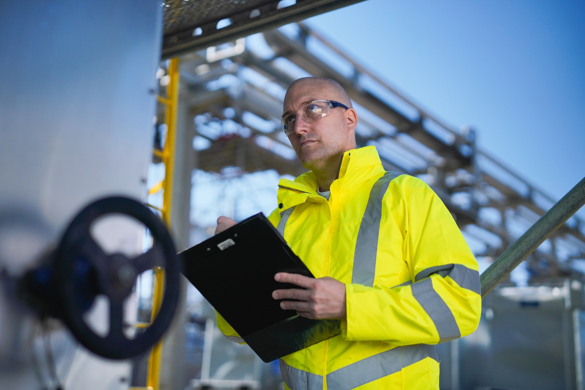 Een kale man in een opvallende gele jas en veiligheidsbril staat buiten bij industriële leidingen. Hij houdt een klembord vast en kijkt naar voren.