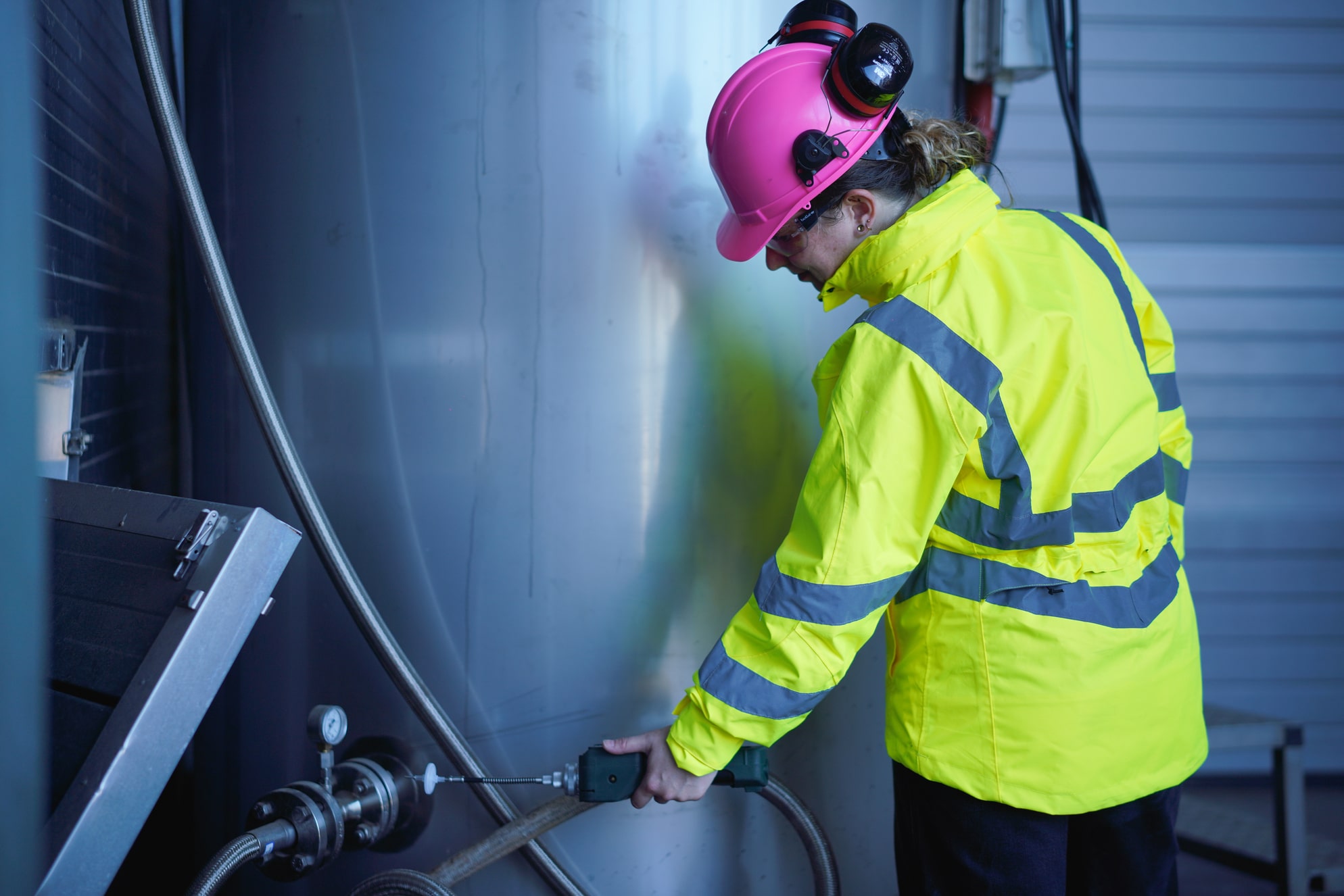 Een technicus in een felgele jas en een roze helm staat voor een grote metalen tank. Ze is bezig met het bedienen van een apparaat dat aan de tank is gekoppeld en draagt veiligheidsoorbeschermers.