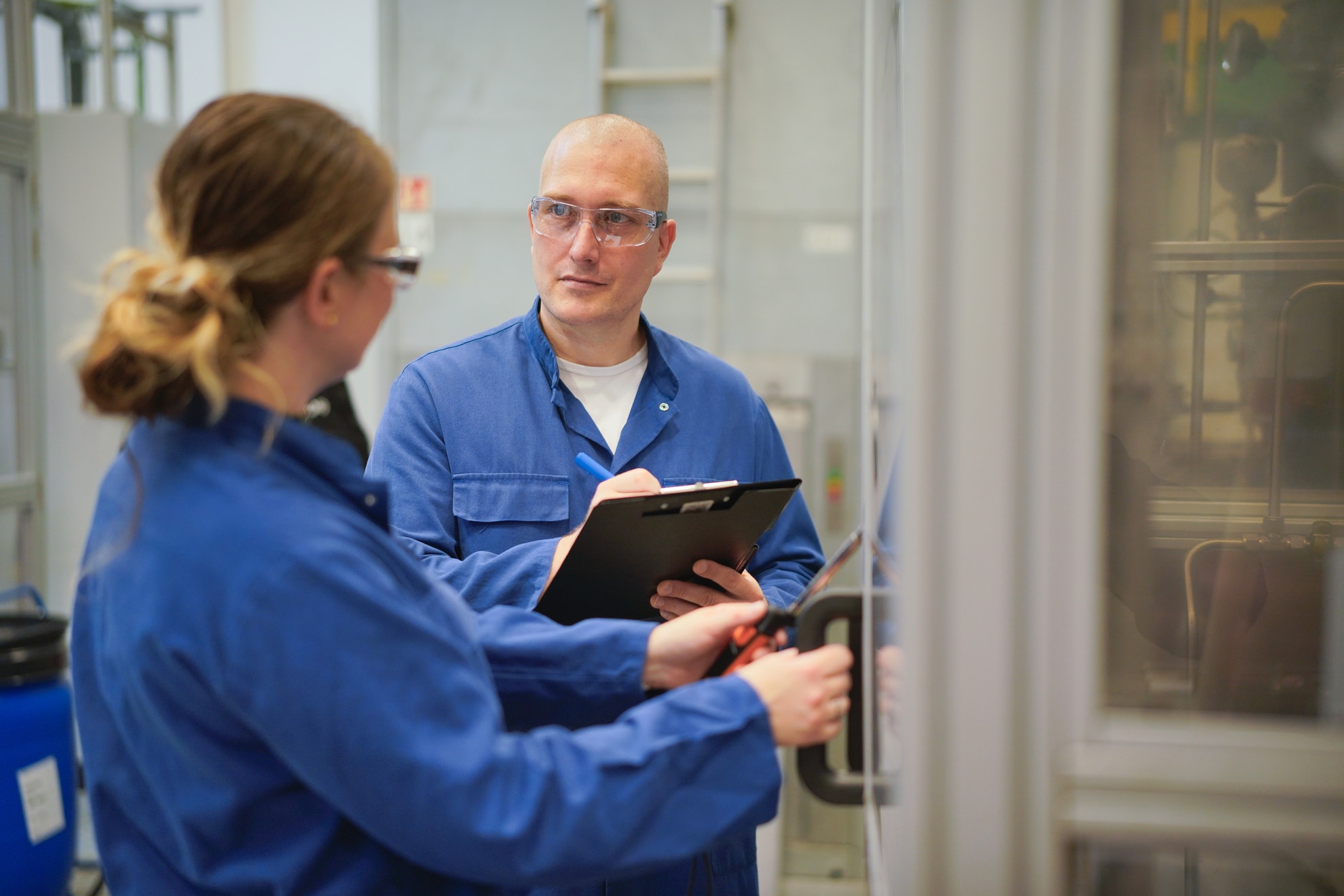Twee technici, gekleed in blauwe overalls en met veiligheidsbrillen op, staan in een werkplaats. Een man met een notitiebord in zijn handen kijkt naar een vrouw die een deur vasthoudt.