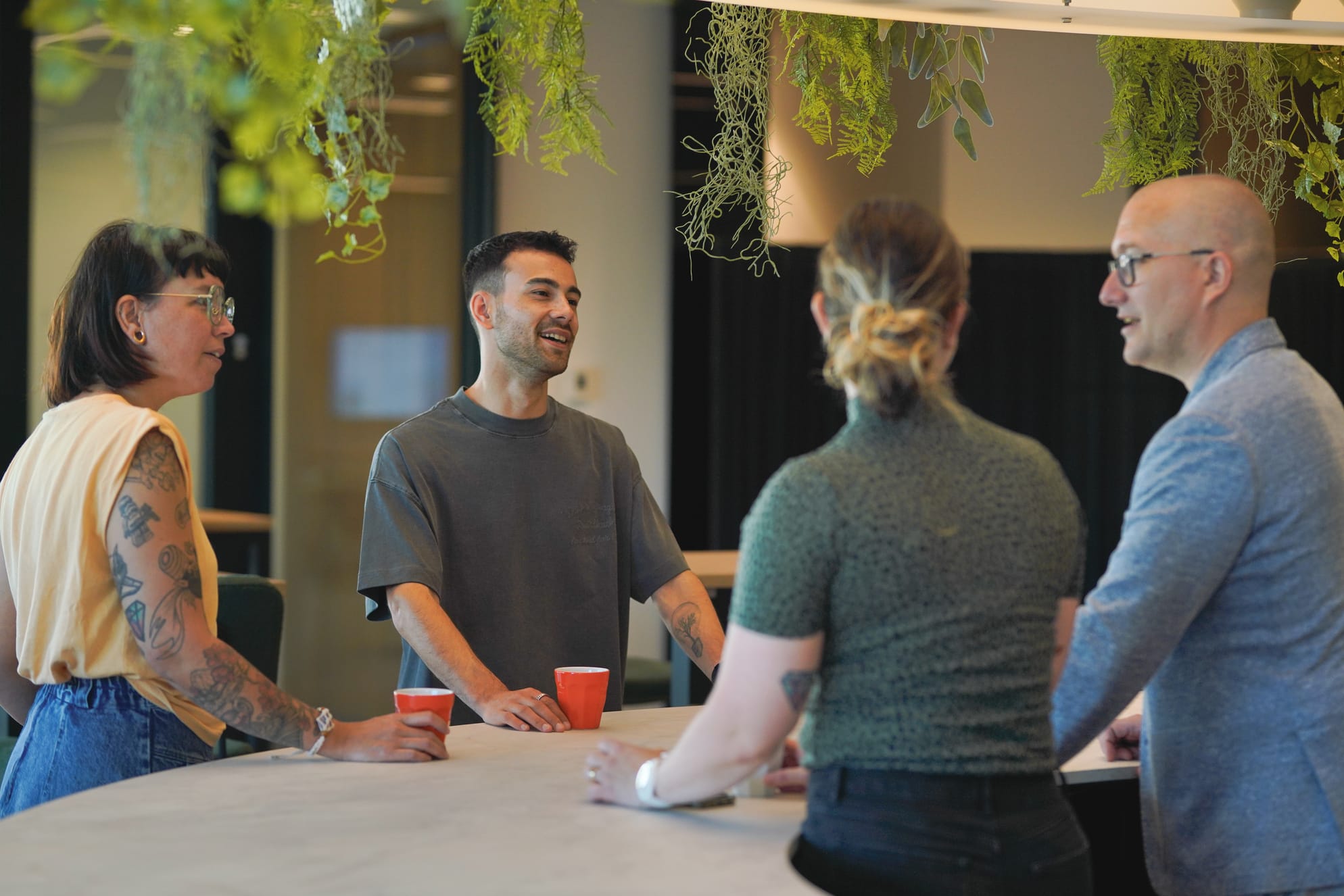 Vier collega's staan rond een tafel in een modern kantoor. Een man in het midden van de foto lacht en praat met zijn collega's. Op de tafel staan twee rode bekers. Boven de tafel hangen planten.
